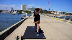 Male fitness model in lunge position on the Rolley exercise mat. Scene shows large 2m x 1m size with full range of movement. Dumbells and skipping rope are sitting beside the thick exercise mat.